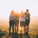 four people standing with their arms around each other looking at the sunset