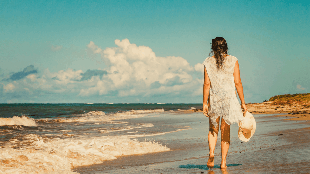 woman in white coverup walking down the beach with hat in hand