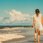 woman in white coverup walking down the beach with hat in hand