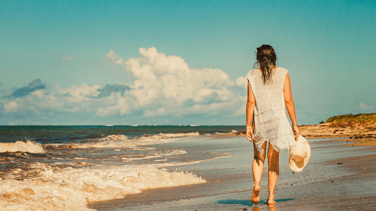 woman in white coverup walking down the beach with hat in hand